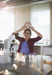Portrait playful girl student holding molecular structure on head at microscope in laboratory classroom - CAIF26569