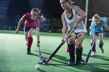 Junge Feldhockeyspielerinnen laufen zum Ball und spielen Feldhockey auf einem nächtlichen Feld - CAIF26629