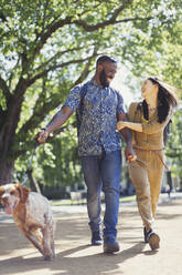 Lächelndes, glückliches junges Paar beim Spaziergang mit dem Hund in einem sonnigen Park - CAIF26647