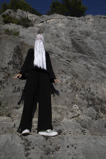 Woman dressed in black wearing crocheted white headdress with fringes standing in front of rock face - PSTF00691
