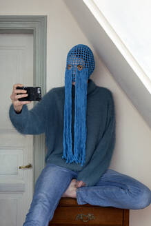 Portrait of young woman wearing crochetedblue headdress with fringes taking selfie with smartphone - PSTF00713