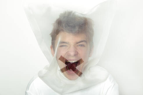 Portrait of screaming teenage boy wearing transparent mask with cross on his mouth - PSTF00737