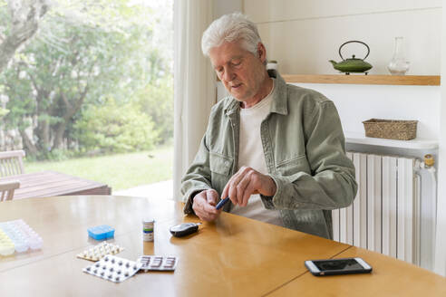 Älterer Diabetiker im Ruhestand, der am Tisch sitzend ein Glaukomgerät zur Blutzuckermessung benutzt - AFVF06165