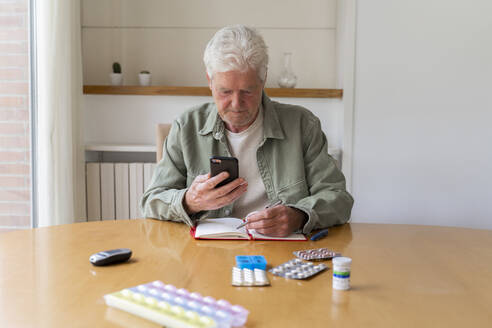 Ein älterer Mann benutzt sein Smartphone, während er zu Hause mit einem Tagebuch und Medikamenten auf dem Tisch sitzt - AFVF06167