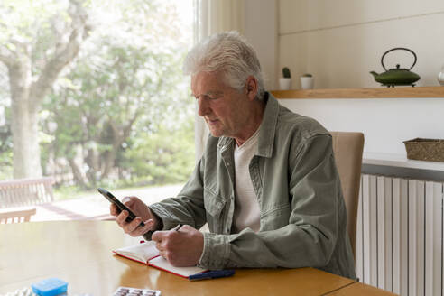 Älterer Diabetiker, der ein Mobiltelefon benutzt, während er zu Hause in sein Tagebuch schreibt - AFVF06169