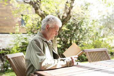 Seitenansicht eines Mannes im Ruhestand, der einen Roman liest, während er an einem Holztisch im Hinterhof an einem sonnigen Tag sitzt - AFVF06180