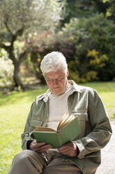 Senior man reading book while sitting in back yard on sunny day - AFVF06182