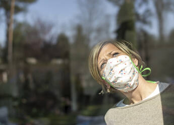 Woman with face mask looking through window glass on sunny day during quarantine - BFRF02222