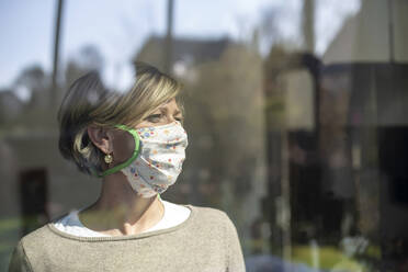 Woman wearing floral mask while looking away through window during isolation - BFRF02224