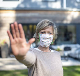 Portrait of woman wearing protective face mask while standing with stop gesture against house during isolation - BFRF02230