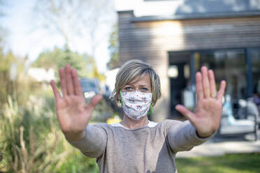 Portrait of mature woman wearing protective face mask while standing with stop gesture against house during epidemic lockdown - BFRF02231