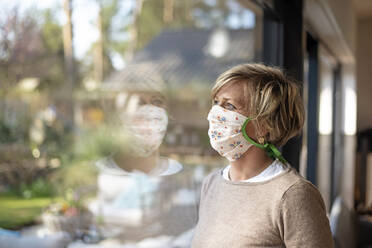 Mature woman wearing floral mask while looking through window at home during COVID-19 isolation - BFRF02232