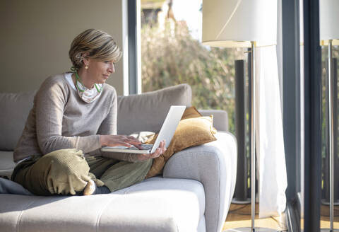 Woman using laptop while sitting on sofa in living room during working from home - BFRF02233