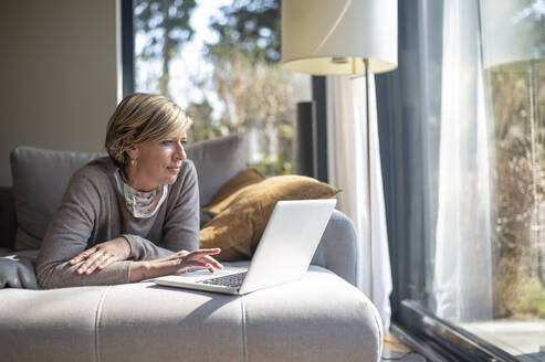 Reife Frau, die einen Laptop benutzt, während sie auf dem Sofa im Wohnzimmer liegt und von zu Hause aus arbeitet - BFRF02234