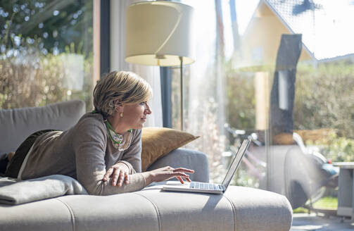 Frau auf dem Sofa liegend mit Laptop im Wohnzimmer bei der Arbeit zu Hause während der Selbstquarantäne - BFRF02235