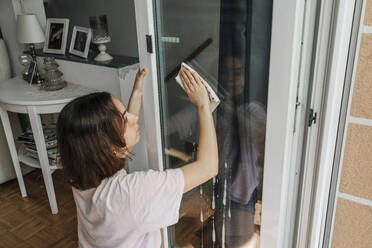 Teenage girl cleaning glass pane of balcony door - OGF00299