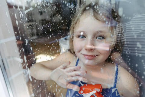 Porträt eines lächelnden blonden Mädchens mit Sprühflasche, das durch ein nasses Fenster schaut, lizenzfreies Stockfoto