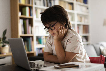 Cute boy with hand on chin looking at laptop while sitting in living room - VABF02876