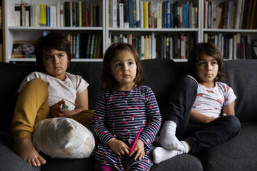 Cute siblings watching TV while sitting on sofa against bookshelf in living room at home - VABF02880