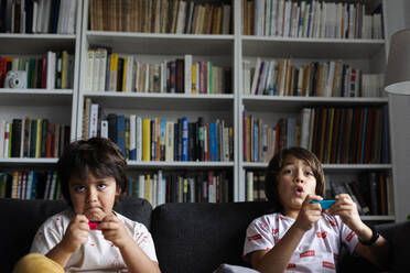 Boys playing video game while sitting on sofa against bookshelf in living room at home - VABF02884