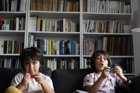 Jungen spielen ein Videospiel, während sie auf dem Sofa vor dem Bücherregal im Wohnzimmer sitzen - VABF02884