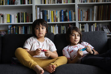 Brothers playing video game while sitting on sofa against bookshelf in living room at home - VABF02885