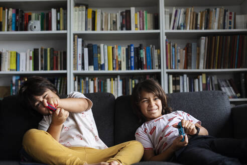 Siblings playing video game while sitting on sofa against bookshelf in living room at home - VABF02886