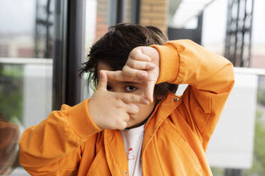 Nahaufnahme eines Jungen, der durch seine Hände schaut, während er am Fenster steht - VABF02891