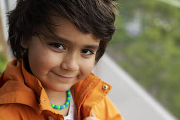 Nahaufnahme des Porträts eines lächelnden Jungen im Grundschulalter, der eine orangefarbene Jacke trägt und im Hinterhof steht - VABF02892