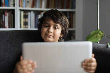 Portrait of boy holding laptop while sitting on sofa against bookshelf in living room at home - VABF02894