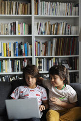 Jungen sitzen mit Laptop auf Sofa gegen Bücherregal im Wohnzimmer zu Hause - VABF02898
