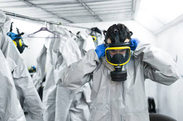 Sanitärarbeiter trägt eine Gasmaske, während er in einem Umkleideraum mit Schutzanzügen steht - JCMF00658