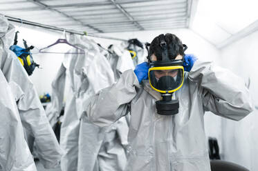 Sanitation worker wearing gas mask while standing by protective coveralls in locker room - JCMF00658