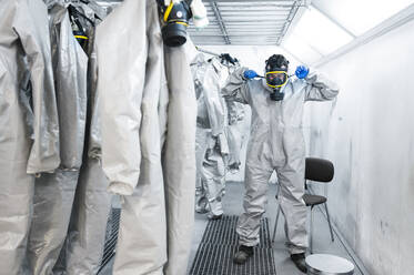 Full length of healthcare worker wearing gas mask while standing by protective coveralls in locker room - JCMF00659