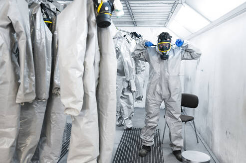 Gesundheitspfleger mit Gasmaske in voller Länge, während er in einem Umkleideraum mit Schutzanzügen steht - JCMF00659