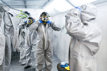 Team of sanitation workers wearing protective coveralls and gas masks while standing in locker room - JCMF00661