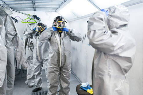 Ein Team von Sanitärarbeitern trägt Schutzanzüge und Gasmasken, während sie in einer Umkleidekabine stehen - JCMF00661