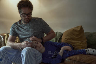 Mann hält Thermometer auf die Stirn seiner kranken Tochter im heimischen Wohnzimmer - ERRF03643