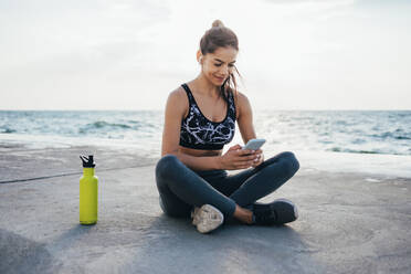 Lächelnde Sportlerin in voller Länge bei der Nutzung eines Smartphones auf der Promenade - OYF00133