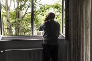 Rückansicht eines älteren Mannes am Telefon, der am offenen Fenster steht - UUF20222