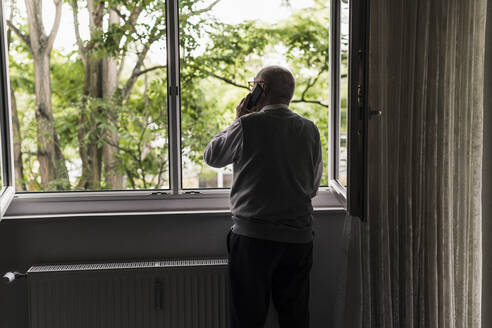 Back view of senior man on the phone standing at open window - UUF20222