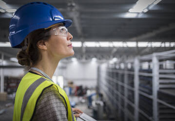 Lächelnde, zuversichtliche weibliche Aufsichtsperson mit Schutzhelm, die in einer Fabrik nach oben schaut - CAIF26738