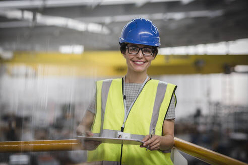 Porträt einer lächelnden, selbstbewussten Arbeiterin mit Klemmbrett auf einer Plattform in einer Fabrik - CAIF26746