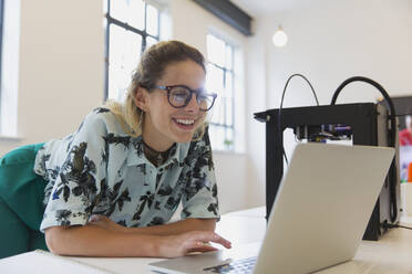 Smiling female designer working at laptop next to 3D printer in office - CAIF26885