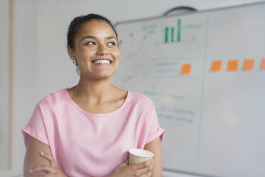 Porträt einer lächelnden, ehrgeizigen Geschäftsfrau, die am Whiteboard im Büro Kaffee trinkt - CAIF26892