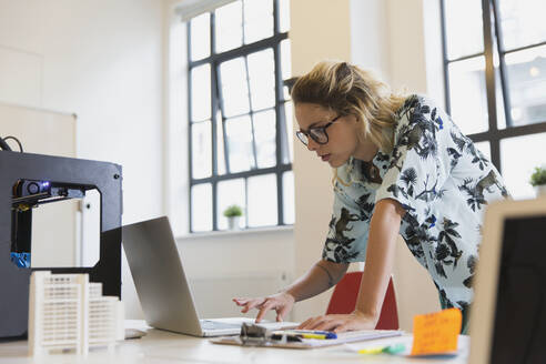 Eine Designerin arbeitet an einem Laptop neben einem 3D-Drucker im Büro - CAIF26899