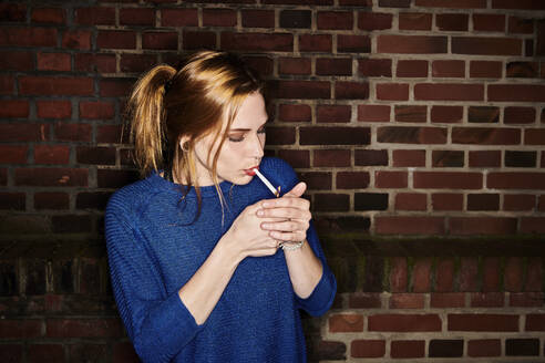 Young woman lighting cigarette against brick wall at night - MMIF00257