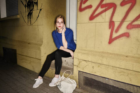 Young woman talking on mobile phone while sitting on steps at night in city stock photo