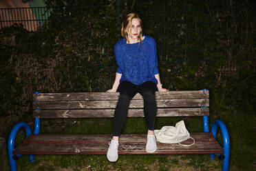 Portrait of young woman sitting on bench in park at night - MMIF00263