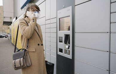 Frau am Telefon mit Gesichtsmaske und Handschuhen in der Nähe eines Paketterminals - AHSF02447